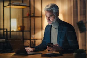 A mature businessman working on laptop at desk indoors in office at night.