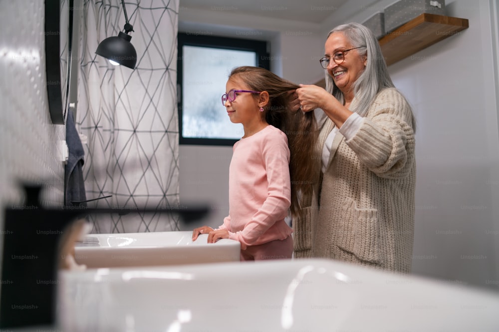 A senior grandmother and granddaughter standing indoors in bathroom, daily routine concept.