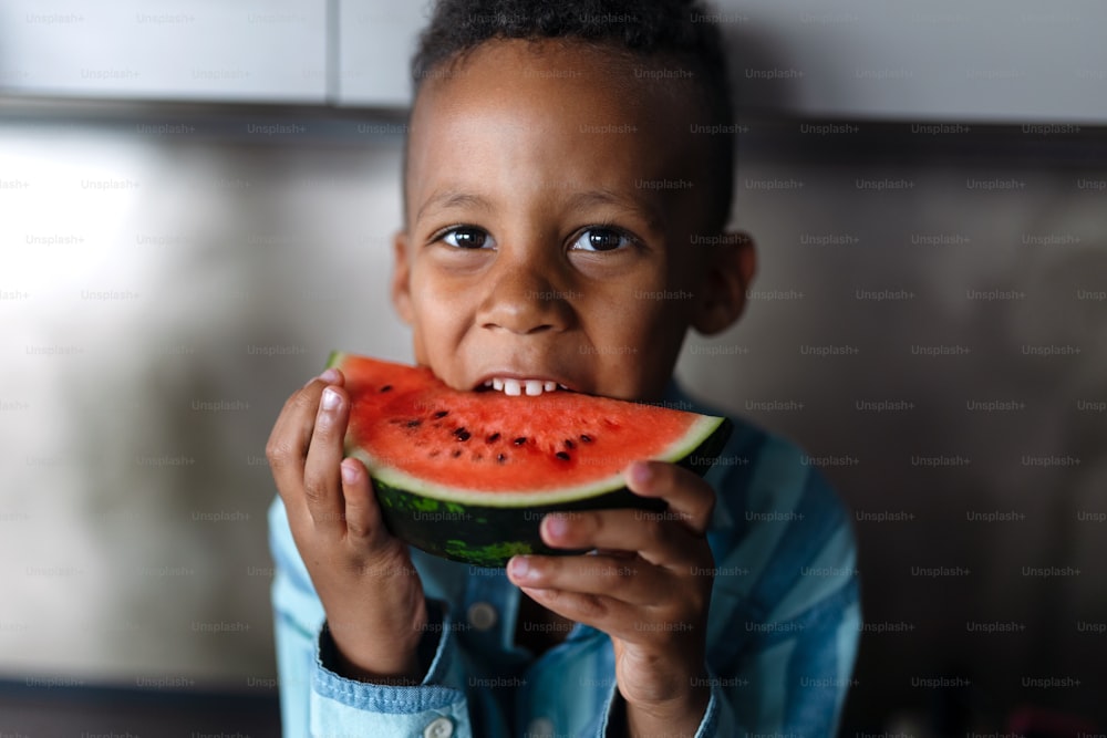 Niño multirracial comiendo un melón en la cocina durante los días calurosos y soleados.
