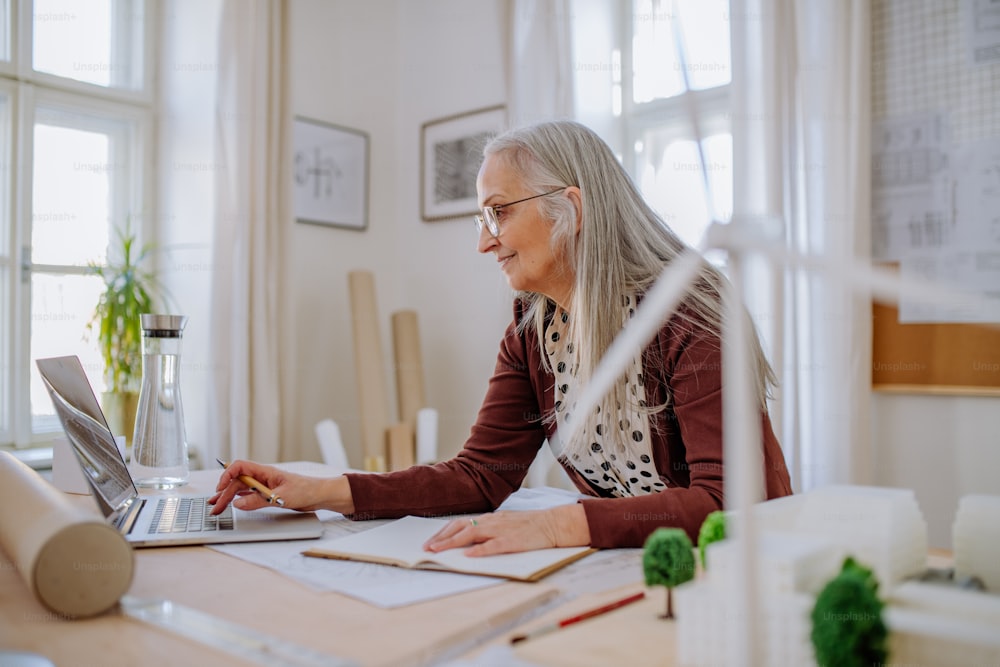 A senior woman architect with model of houses working on laptop in office.