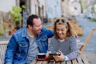 A portrait of happy young man with Down syndrome with his mother sitting in cafe outdoors and talking.