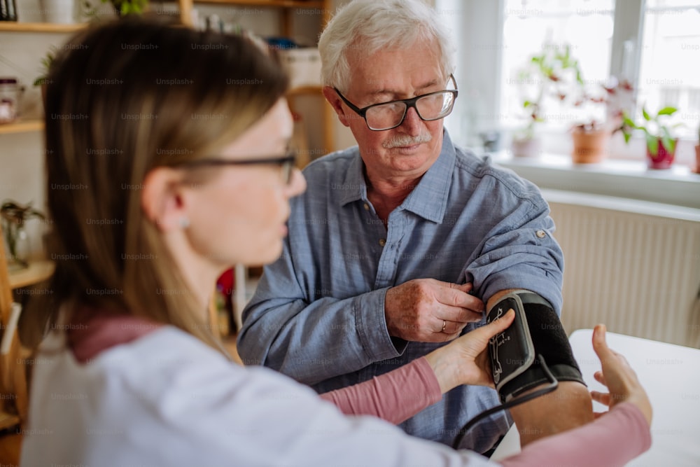 A female doctor visiting senior man and examinig him indoors at home, measuring blood pressure.