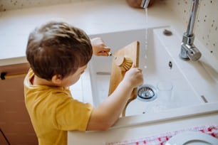 A little boy washing cup in sink in kitchen with wooden scrub, sustainable lifestlye.