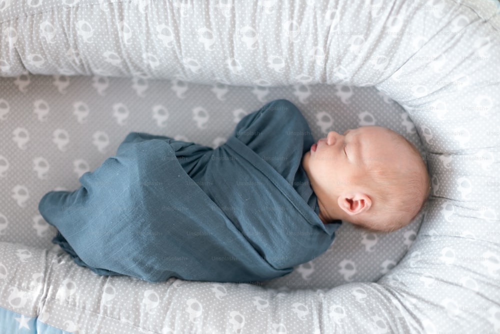 Um bebê recém-nascido dormindo e envolto em pano azul deitado em ninho cinza.