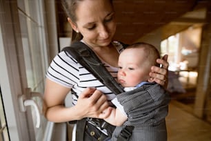 Beautiful young mother at home with her cute little son in baby carrier.