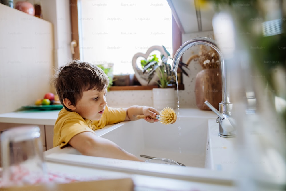 小さな男の子がキッチンのシンクでコップを洗う、木製のスクラブ、持続可能な生活。
