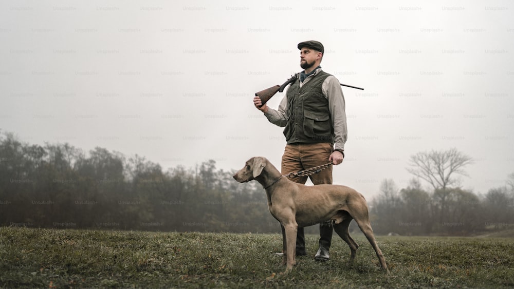 Un cazador con perro en ropa de tiro tradicional en el campo sosteniendo una escopeta.