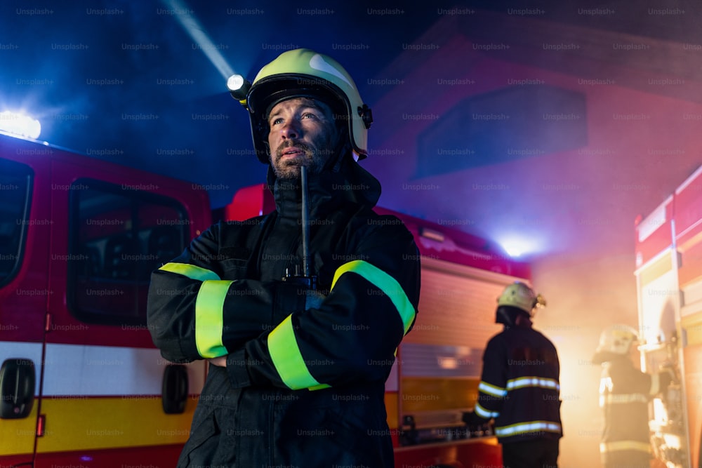 Una vista de ángulo bajo de un bombero hablando con un walkie talkie con un camión de bomberos en el fondo por la noche.