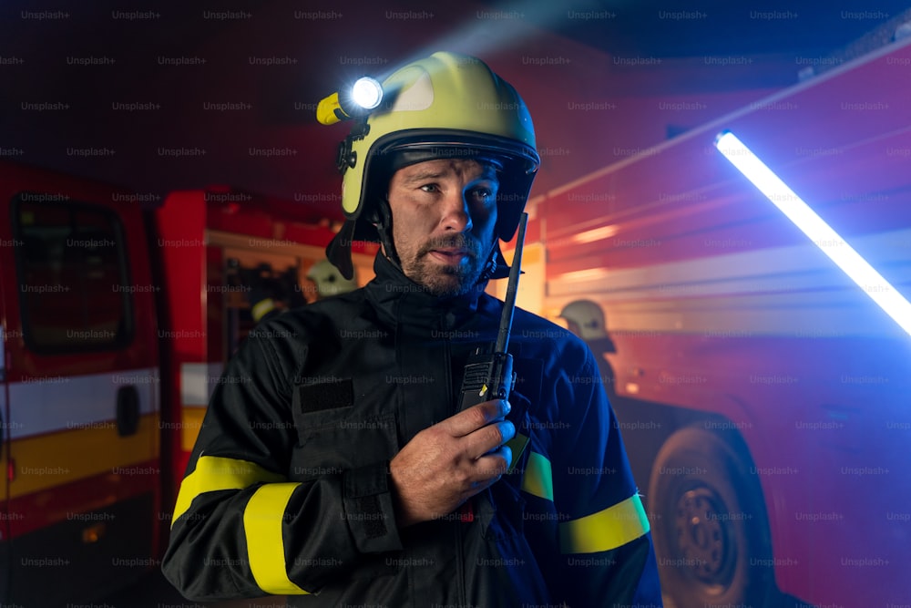 A firefighter talking to walkie talkie with fire truck in background at night.