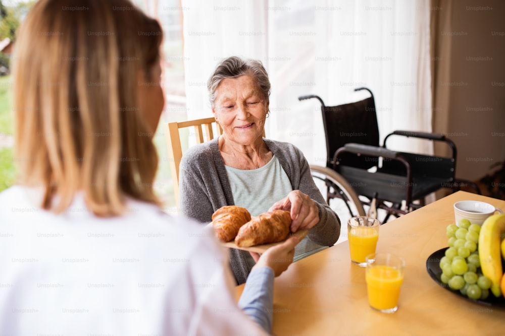 訪問中の健康訪問者と高齢の女性。テーブルに座る年配の女性に食事を与える看護師。
