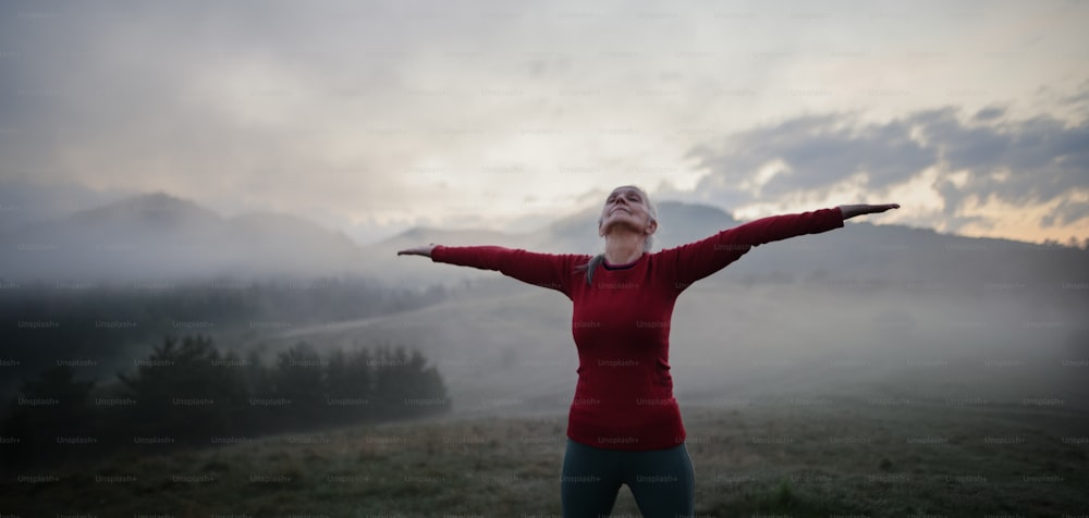 Uma mulher idosa fazendo exercícios de respiração na natureza no início da manhã com neblina e montanhas ao fundo.