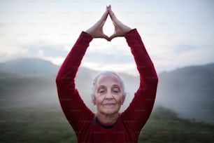 A senior woman doing breathing exercise in nature on early morning with fog and mountains in background.