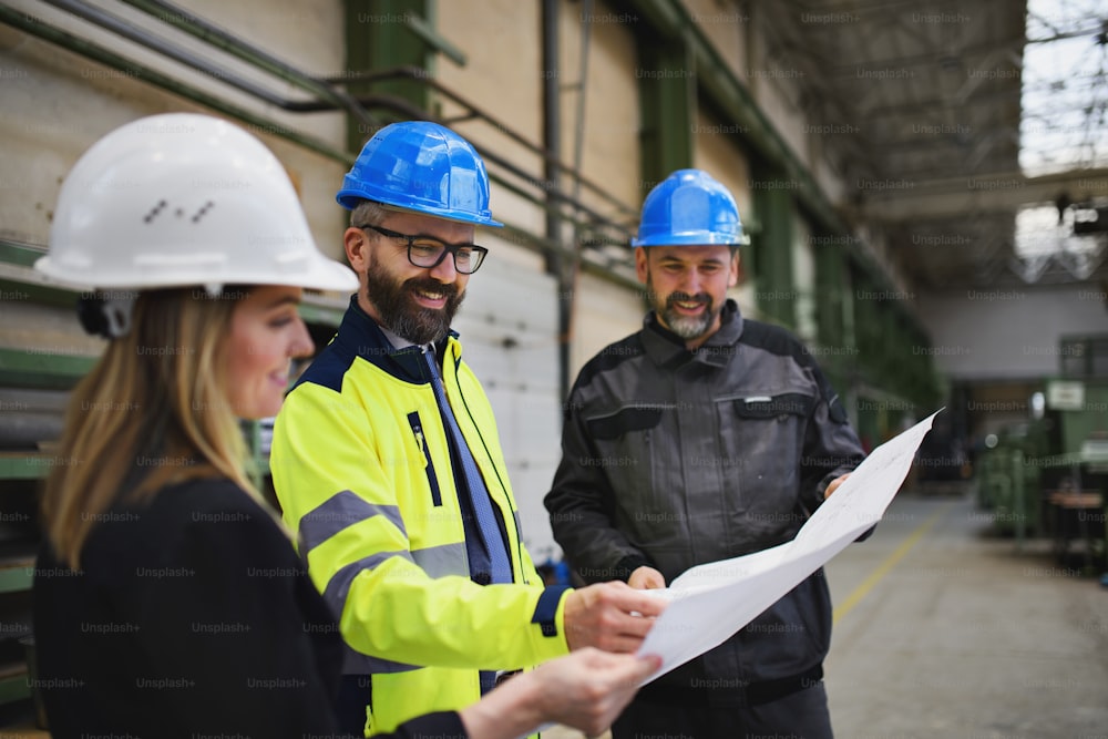 Um gerente supervisores, engenheiro e trabalhador industrial de uniforme discutindo plantas em grande salão de fábrica de metal.
