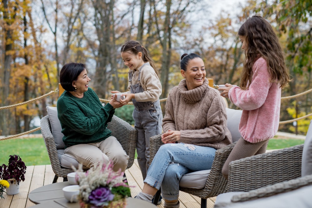 Two happy sisters with a mother and grandmother sitting and drinking tea outdoors in patio in autumn.