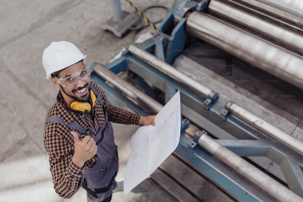 A high angle view of heavy industry worker with safety headphones and hard hat in industrial factory holding blueprints