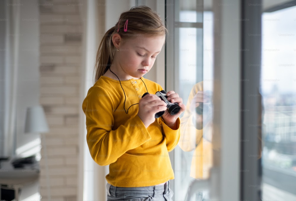 A curious little girl with Down syndrome with binoculars looking through on window at home.