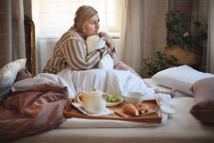 Una mujer feliz con sobrepeso con auriculares y teléfono inteligente desayunando en la cama en casa.