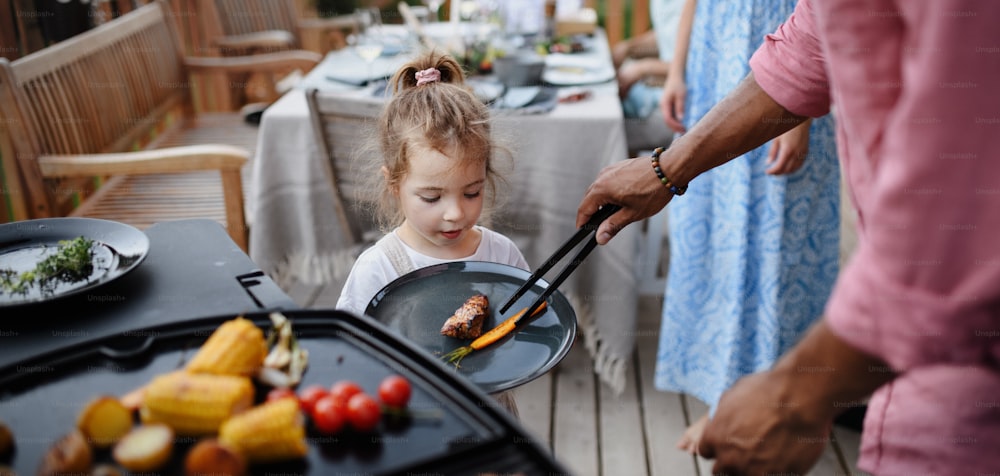 Un padre poniendo carne y verduras a la parrilla en el plato a su hija durante la fiesta familiar de verano en el jardín.
