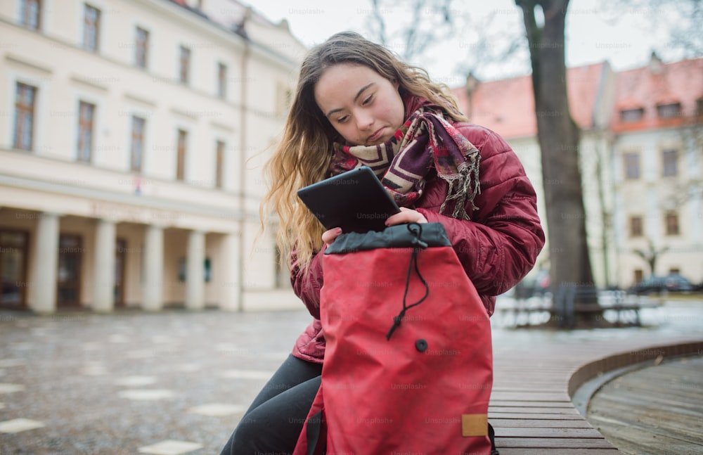 A young woman with Down syndrome sitting on bench and using tablet in town in winter