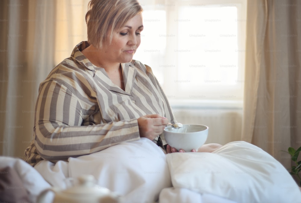 Una mujer feliz con sobrepeso con auriculares y teléfono inteligente desayunando en la cama en casa.