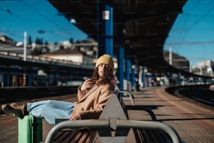 짐을 들고 기차역 플랫폼에 혼자 앉아 있는 젊은 여행자 여성.