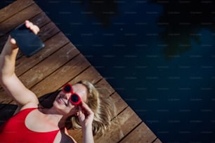 A rlaxed woman wearing headphones listening to music and taking selfie when lying on a pier by natural lake in summer