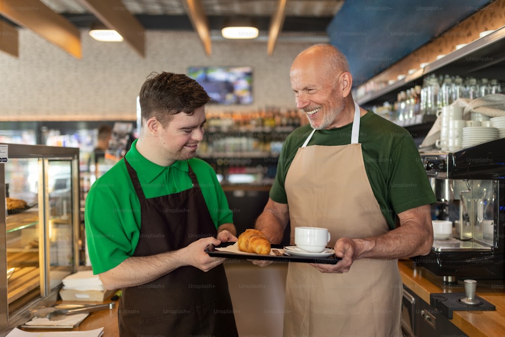 다운 증후군을 앓고있는 행복한 웨이터가 카페에서 동료의 도움으로 커피를 제공합니다.