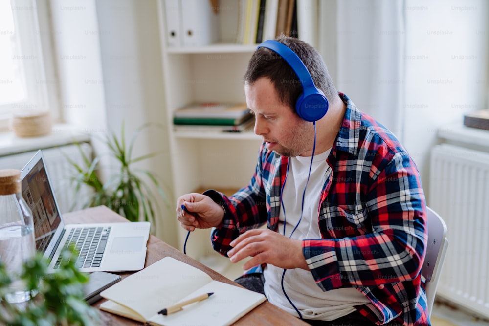 A young man with Down syndrome sitting at desk in office and using laptop, listening to music from headphones.