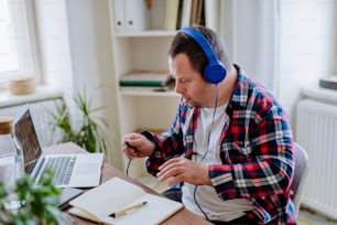 A young man with Down syndrome sitting at desk in office and using laptop, listening to music from headphones.