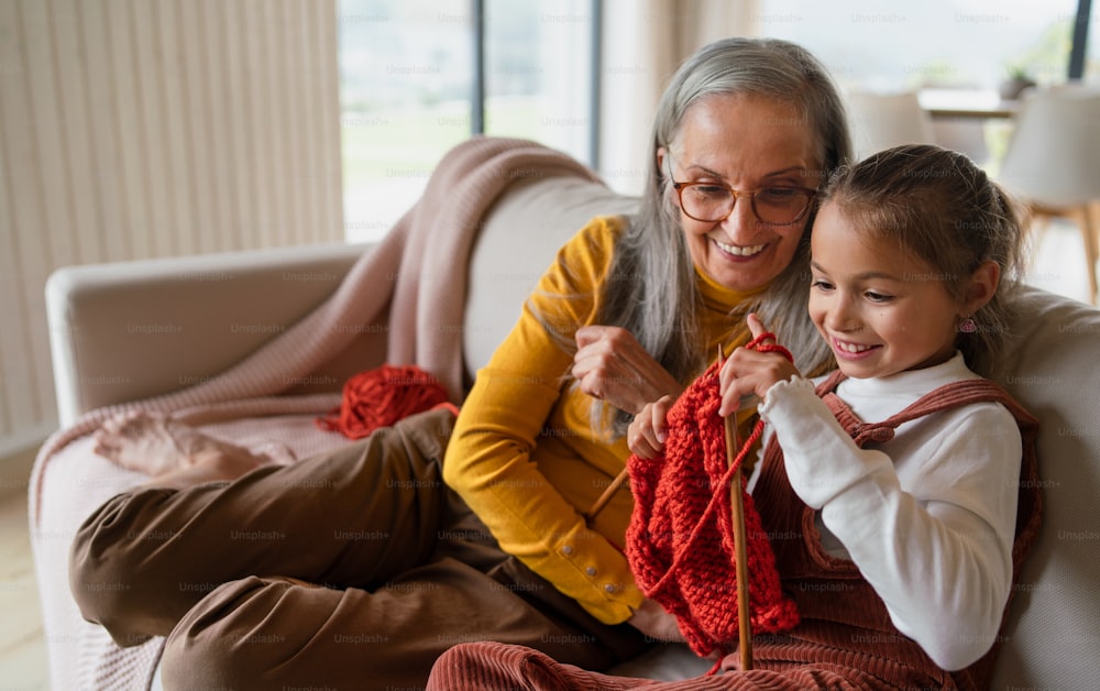 A little girl sitting on sofa with her grandmother and learning to knit indoors at home.