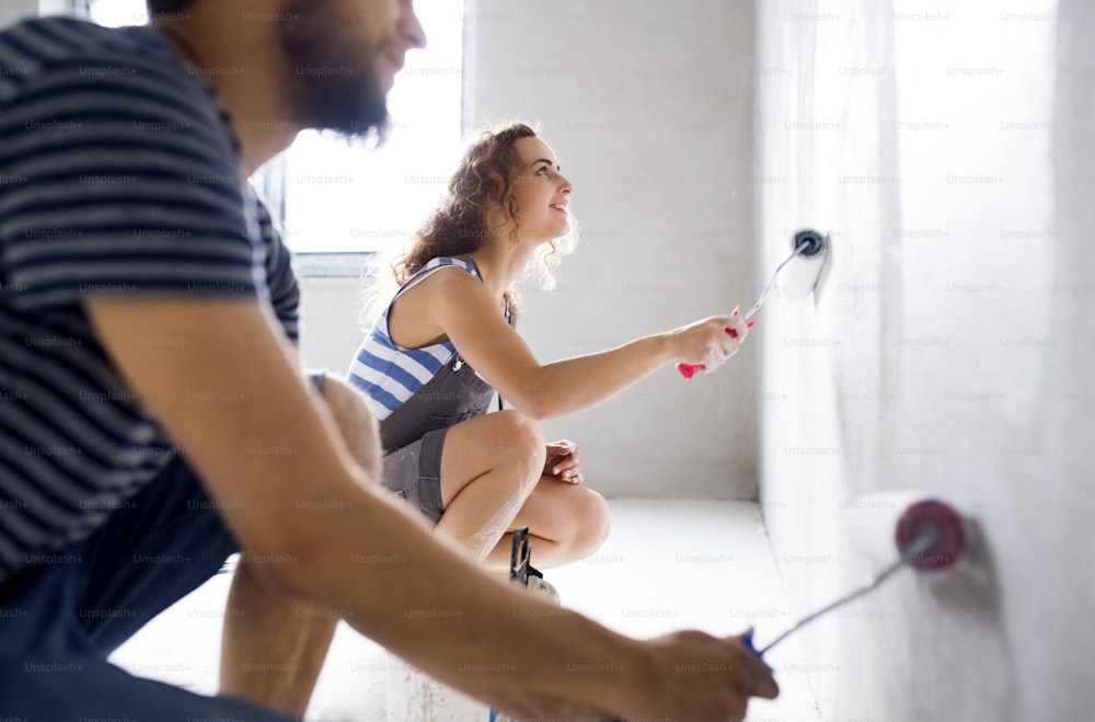 Beautiful young couple having fun and painting walls using paint rollers in their new house. Home makeover and renovation concept.