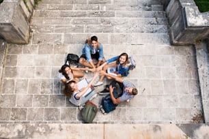 Grupo de estudantes adolescentes atraentes sentados em degraus de pedra em frente à universidade, empilhando as mãos.