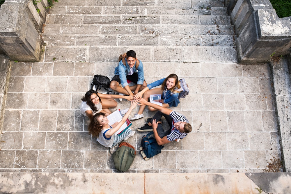 Group of attractive teenage students sitting on stone steps in front of university, stacking hands.