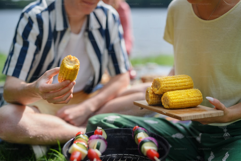 A close-up of young friends putting corn on grill and having barbecue when camping in campground.