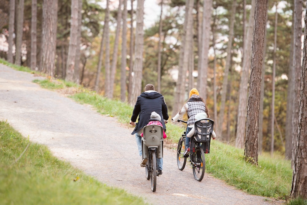 Beautiful young family with two daughters in bicycle seats in warm clothes cycling outside in autumn nature. Rear view.