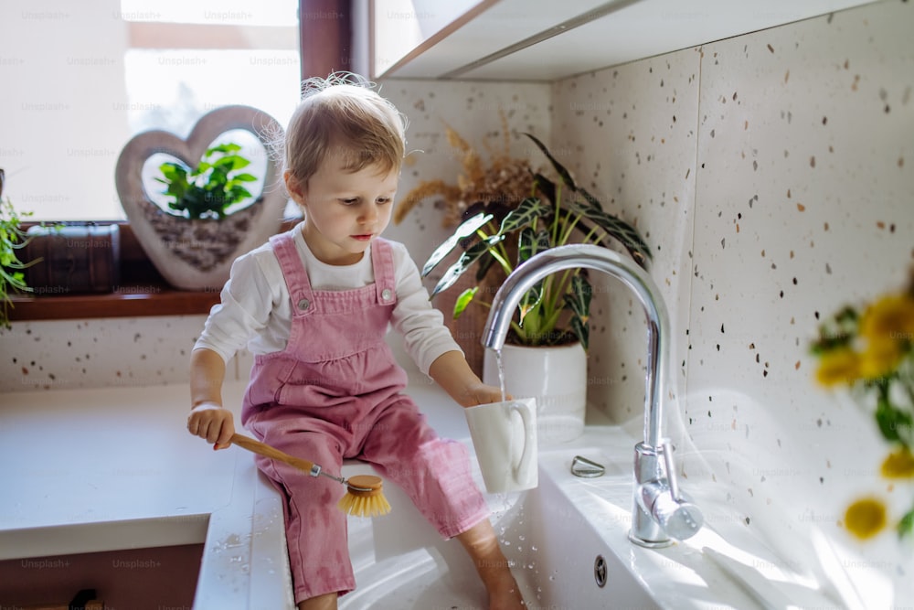 Una niña sentada en la encimera de la cocina y lavando la taza en el fregadero de la cocina.
