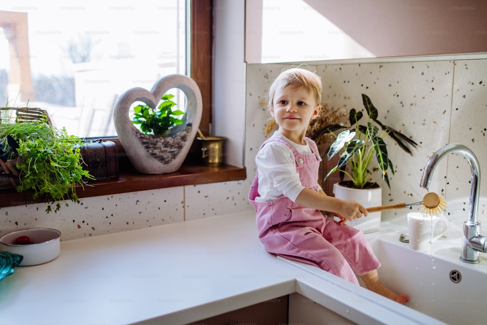 Una niña sentada en la encimera de la cocina y lavando la taza en el fregadero de la cocina.