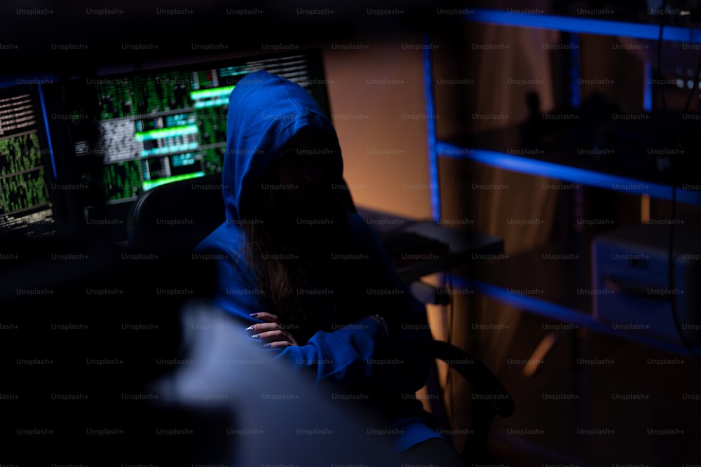 A hooded anonymous hacker by computer in the dark room at night, cyberwar concept.