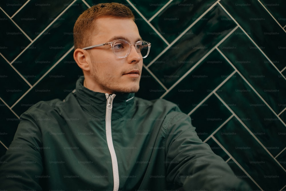 A portrait of young man in jacket and glasses sitting agiant dark tiled wall.