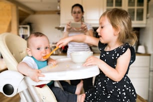 Two little children at home, adorable toddler girl feeding her baby brother using spoon. Their mother taking picture with smart phone.