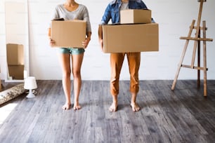 Unrecognizable young married couple moving in new house, holding big cardboard boxes.