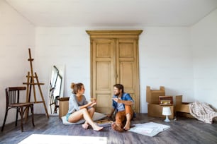 Young married couple moving in new house, sitting on the floor near cardboard boxes, eating pizza.