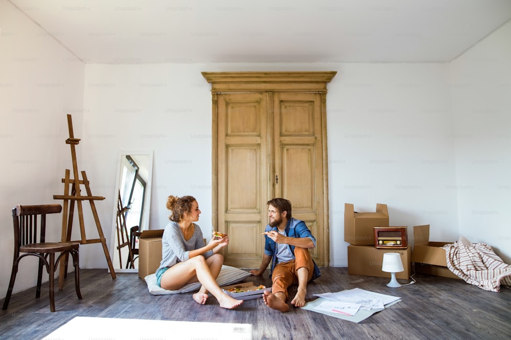 Young married couple moving in new house, sitting on the floor near cardboard boxes, eating pizza.