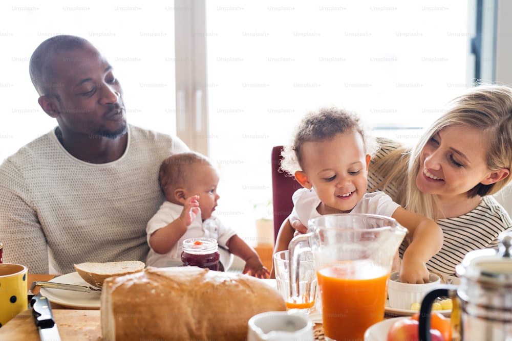 Hermosa familia joven interracial en casa con su linda hija y su pequeño bebé desayunando juntos.