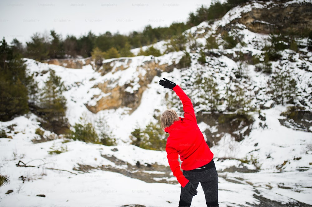 Femme âgée faisant du jogging à l’extérieur dans la nature hivernale, s’étirant. Vue arrière.