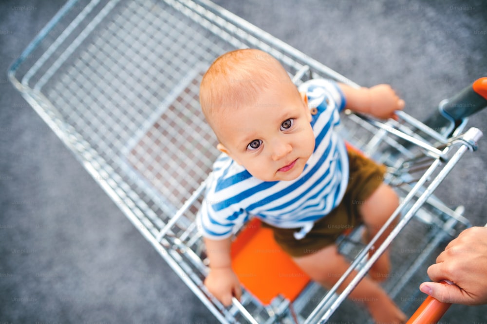 Baby boy with unrecognizable mother sitting in the shopping trolley outside. Top view
