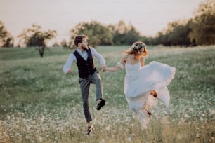 Beautiful young bride and groom outside in green nature at romantic sunset, holding hands, jumping high.