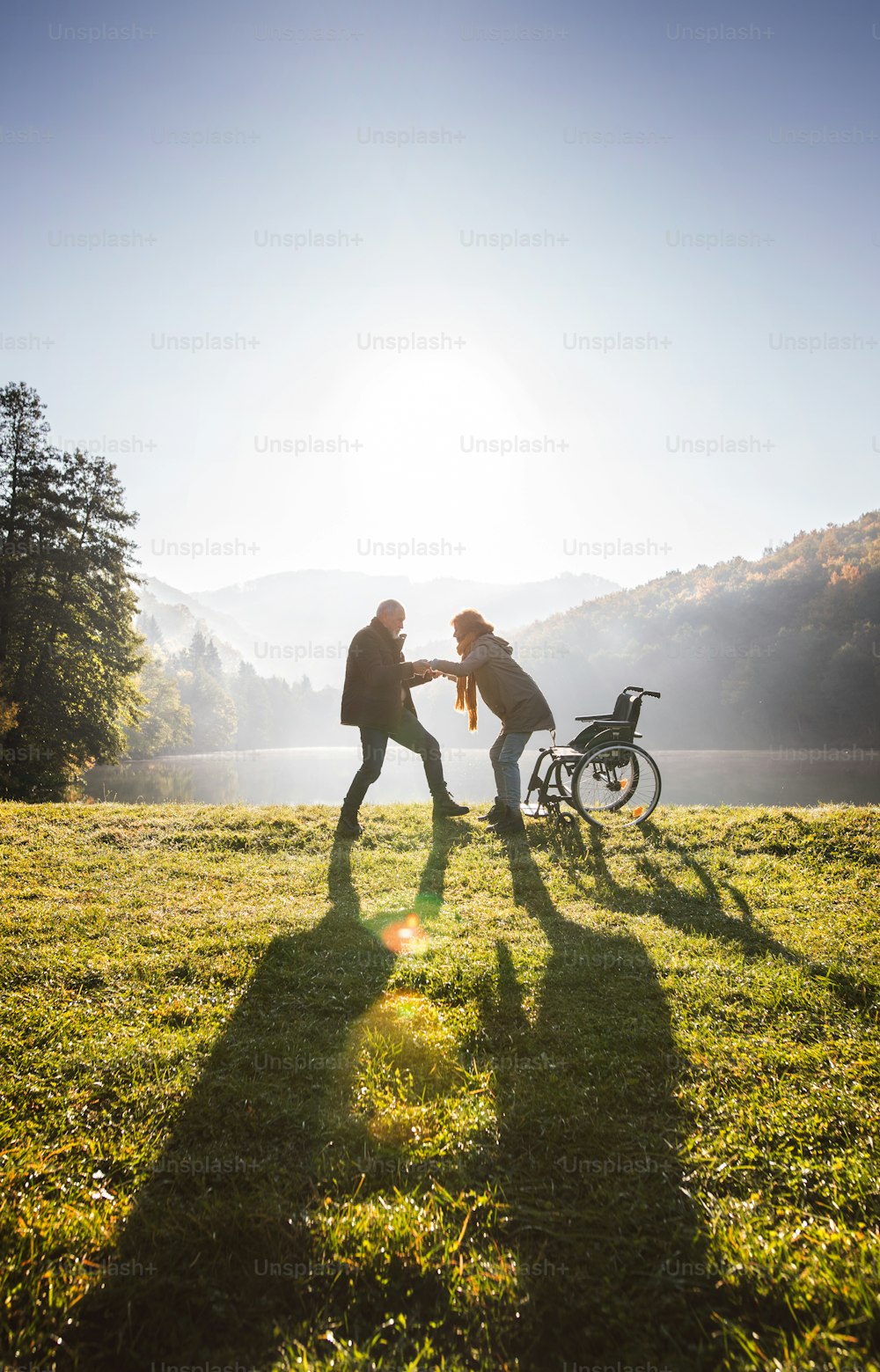 秋の美しい自然の中を散歩するアクティブなシニアカップル。早朝、湖畔で車椅子に乗った男女。
