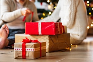 Couple de personnes âgées méconnaissables assis sur le sol devant un arbre de Noël illuminé à l’intérieur de leur maison se donnant des cadeaux.