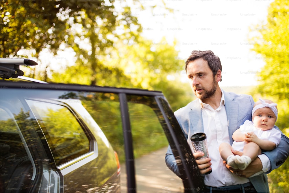 Young father carrying his baby girl. Man at the car, holding takeaway coffee.